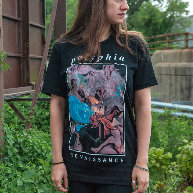 Black short sleeve shirt with polyphia written in white font across the top of the chest. Below that, there is a distorted colorful renaissance painting, Below the painting there is smaller white text that says RENAISSANCE. An individual is modeling the shirt and standing in front of green bushed and a rusted overpass.