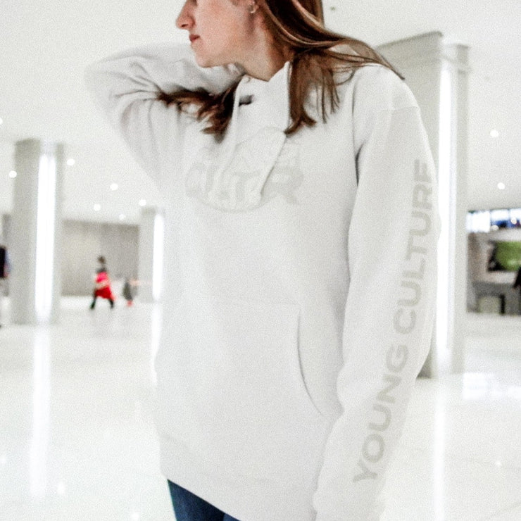 White pullover hoodie with "YNG CLTR" written on the chest in grey lettering, inside a grey globe. "YOUNG CULTURE" is written vertically along either arm in grey font. An individual is wearing the hoodie in a white indoor area with pillars in the background.