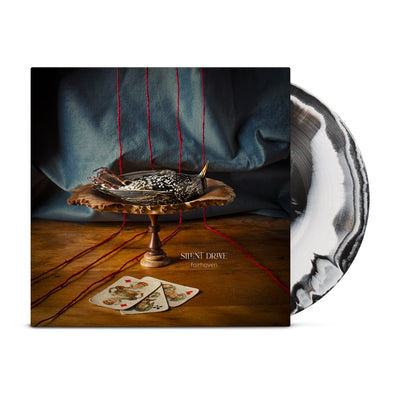 An image of a square LP cover with vinyl disc half exposed. LP cover features a wooden centerpiece positioned on a wooden floor with a blue curtain behind. 4 strands of yarn run behind the centerpiece across the floor. On the centerpiece lies a dead bird and at its base lie 3 playing cards. Vinyl Disc is a mixed black and white LP