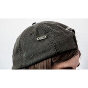 Enamel pin with "COLD" written in silver lettering over a black background. Black nails form the hole inside the O. Pin is being worn on a grey hat.