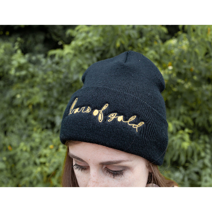 Black beanie with BARS OF GOLD written across the front in yellow cursive. An individual is modeling the beanie and standing in front of greenery.