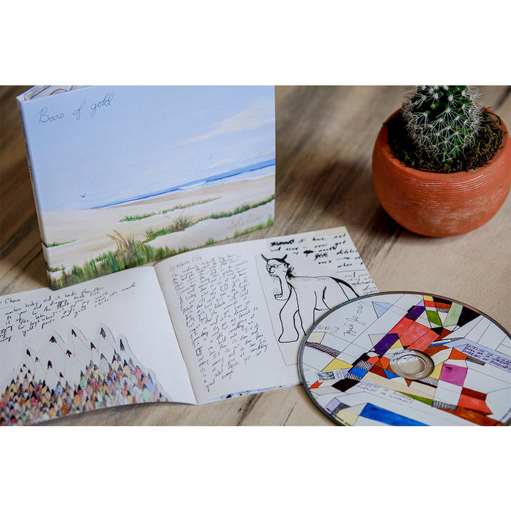 Square CD with a drawing of a beach with tall grass and birds flying in the sky. In the top left corner there is black cursive font that says BARS OF GOLD. There is a colorful CD laying next to the cover. Both are laying on a wooden surface next to a cactus plant.