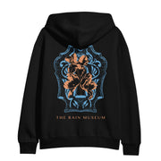  The back of the hoodie is a blue and orange colored crest design with THE RAIN MUSEUM written in orange text below it.