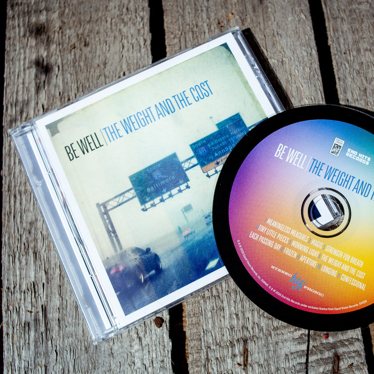 Square CD with black text in the top left that says BE WELL. Next to that there is blue text that says THE WEIGHT AND THE COST. The album artwork is an image of a foggy highway with cars driving and signs above the road. A CD disc is laying on top of the cover and it is rainbow colored. Both are laying on top of a wooden surface.