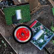 Square CD with text in the top left corner that says CALLING ALL CAPTAINS. In the center there is a white coffin surrounded by trash all over the ground. There is a red CD disc on top of the cover that has a drawing of a rose in the center. There is white texts on the disc that says CALLING ALL CAPTAINS. Both are laying on top of dirt and sprouts coming out of the ground.