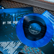 Vinyl jacket with large white text that says BY THE GRACE OF GOD across the center with smaller text below that says PERSPECTIVE. Album artwork is an image of buildings with a dark blue filter over it. peeking out of the jacket is a black and blue colored vinyl. Both are laying on top of metal stairs.