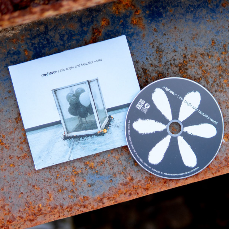 Square CD with text that says greyhaven in black font. Next to that there is more black text that says this bright and beautiful world. The album artwork is a bunch of balloons inside a glass box. Surrounding the box is dirt and some flowers along the edges. Laying next to the cover is a CD disc with a white flower. Both are laying on top of a rusted surface. 