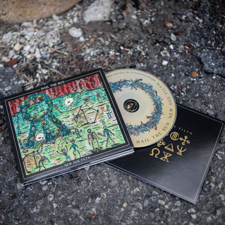 Square CD, the album cover is skeletons surrounding the Hail The Sun logo in front of a forest. Above the drawing there is text that says HAIL THE SUN, and below the drawing there is text that says NEW AGE FIILTH. There is a CD peeking out of the cover that is yellow with HAIL THE SUN and NEW AGE FILTH written along the border. Both are laying on an asphalt surface.