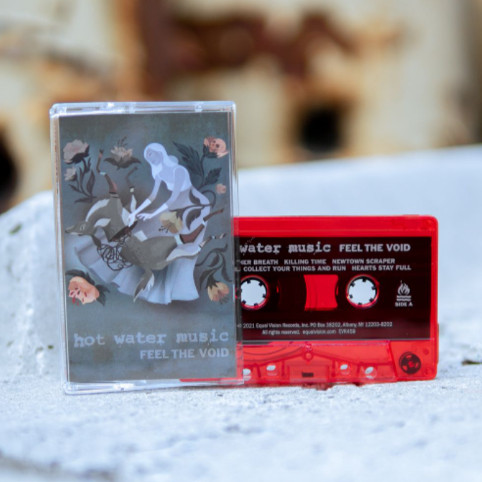 Cassette tape with artwork of a woman wearing a long grey dress in the center. The woman is reaching down onto her legs where there is a deer that has been split in half. In each corner is drawings of light pink flowers, one of them having a skull blooming out of it. On the bottom there is text that lightly says HOT WATER MUSIC and below it there is text that says FEEL THE VOID. To the right is a red cassette with the same text written on it. Both are standing up on a concrete surface.