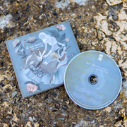Square CD with artwork of a woman wearing a long grey dress in the center. The woman is reaching down onto her legs where there is a deer that has been split in half. In each corner is flowers, one of them having a skull blooming out of it. In the top left corner there is text that says HOT WATER MUSIC and below is text that says FEEL THE VOID. To the right is a disc with the same text written on it, and a drawing of a fire with two lines representing water under it. Both are on top of rocks and dirt.