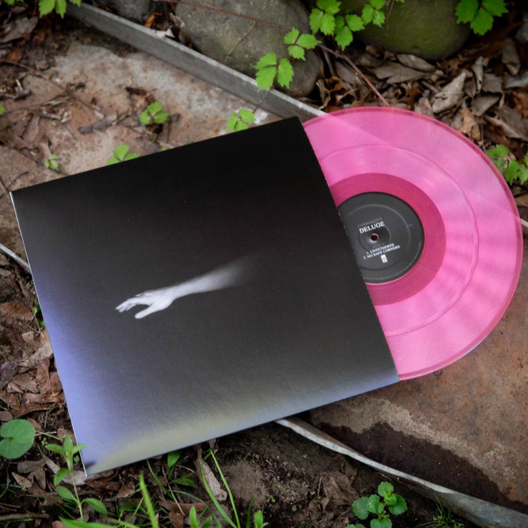 Image of Fairweather Deluge Vinyl LP with LP exposed to show color. Color of LP is Pink. Album cover shows half a ghostly arm on a black background. Both are laying on the ground on dirt and weeds.