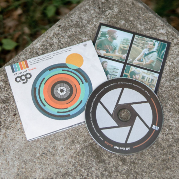 Square CD with artwork of a multicolored camera lens in the center. In the top left there is black lettering that says AGO. Above that is text that says ALL GET OUT, and next to it is orange lettering that says KODAK. To the right is a disc with a design of a camera shutter. Both are laying on top of a granite surface.