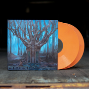 Vinyl jacket with orange / red disc peeking out. Album artwork is a dark forest with one larger tree in the center. The tree has windows built into the trunk with rocks leading up to them. The whole forest is covered in an eerie blue colored fog. In the bottom left, there is text that says "THE DEAR HUNTER", and in the bottom right there is text that says "ACT V HYMNS WITH THE DEVIL IN CONFESSIONAL". The vinyl jacket and discs are on top of planks of wood.