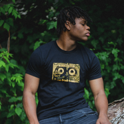 Black short sleeve shirt with orange cassette tape across the chest. On the top of the cassette, there is text that says THE JULIANA THEORY. An individual is modeling the shirt, sitting on a tree trunk in front of green leaves.