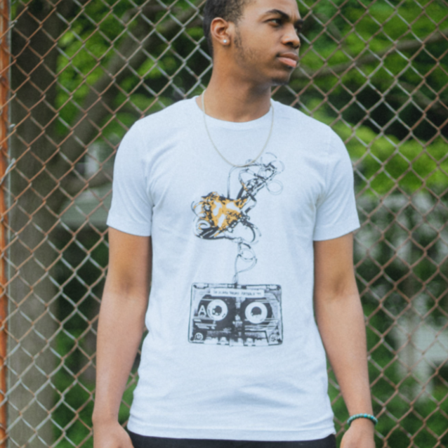 White short sleeve shirt with a drawing of a cassette tape in black and white. Above the cassette tape, the tape is coming out of the top and is all tangled. An individual is modeling the shirt and standing in front of a slightly rusted fence with trees in the background.