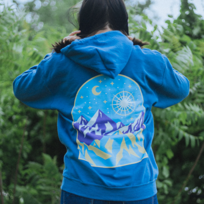 Back of indigo colored hoodie with a drawing of mountains and a night sky. There is a star drawn brightly shining next to the moon above a mountain range. An individual is modeling the shirt standing in front of greenery.