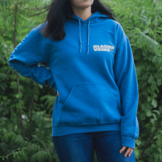 Indigo colored pullover with GLACIER VEINS written in the top corner of the chest in yellow. On the sleeve there is blue text that says LUNAR REFLECTION. An individual is modeling the shirt and standing in front of trees and greenery.