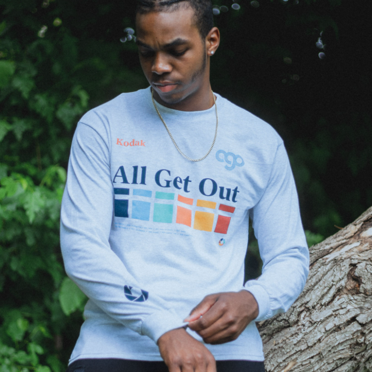 Ash colored long sleeve shirt with text across the chest that says "All Get Out". Below that is a color blocked rainbow design that runs horizontally. In the top corner of the chest, there is orange text that says Kodak. On the bottom of one sleeve there is a camera shutter design. An individual is modeling the shirt, sitting on a tree trunk in front of leaves.