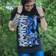 Black short sleeve shirt with Kaonashi written vertically on the left hand side in white font. The background is blue, red, black, and white splattered. There is smaller scratchy text towards the bottom right, also printed in white. An individual is modeling the shirt, standing in front of greenery.