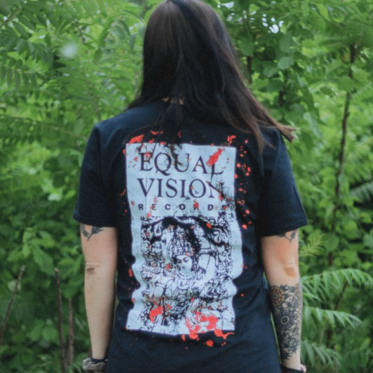Back of black short sleeve shirt with Equal Vision Records logo covering the entirety. Logo printed in white/cream color. Red splatter over logo. An individual is modeling the shirt, standing in front of greenery.