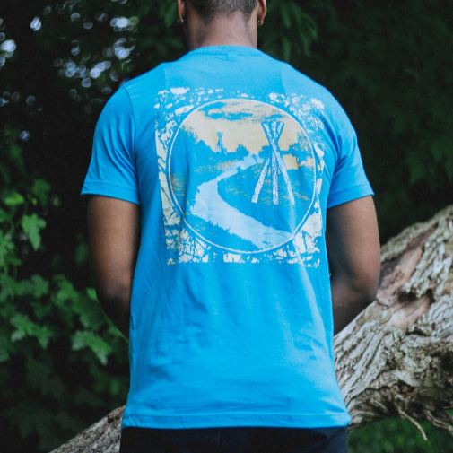Back of short sleeve shirt with a drawing of wooden structures lining a river running into the distance. There is also grass lining the river with trees in the distance. An individual is modeling teh shirt and standing in front of a tree trunk.