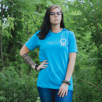 Aqua colored short sleeve shirt with the Hail The Sun logo in top corner (circle with three lines coming out of the bottom). An individual is modeling the shirt and standing in front of trees in a forest.