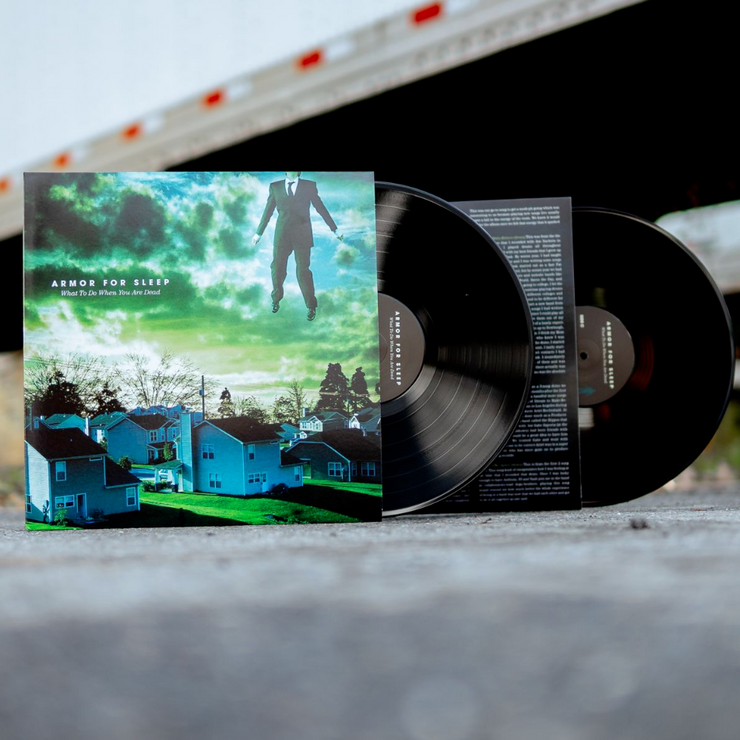 Square vinyl with white text that says ARMOR FOR SLEEP in blue and green tinted clouds. Below that there is smaller text that says WHAT TO DO WHEN YOU ARE DEAD. The album art is an image of a neighborhood of houses. In the top right there is a body of a man wearing a suit, but his head is not in the frame. Two black Records are sticking out of the side.