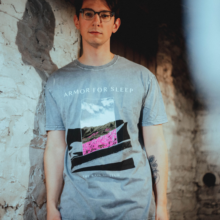 Someone wearing Ash stone colored short sleeve shirt with ARMOR FOR SLEEP written in white text above an image. Below the image there is smaller white text that says THE RAIN MUSEUM. The artwork is black and white hills with a rectangle in the center with hills and pink flowers in the center.