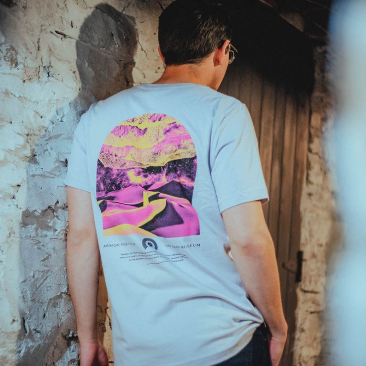 Someone wearing the back of the shirt is artwork of a desert shaded in yellow and pink. Below the artwork there is text that says ARMOR FOR SLEEP and THE RAIN MUSEUM.