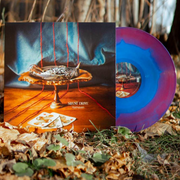 An image of a square LP cover with vinyl disc half exposed. LP cover features a wooden centerpiece positioned on a wooden floor with a blue curtain behind. 4 strands of yarn run behind the centerpiece across the floor. On the centerpiece lies a dead bird and at its base lie 3 playing cards. Vinyl Disc is a blue and red mixed LP. The vinyl is outside during the fall.