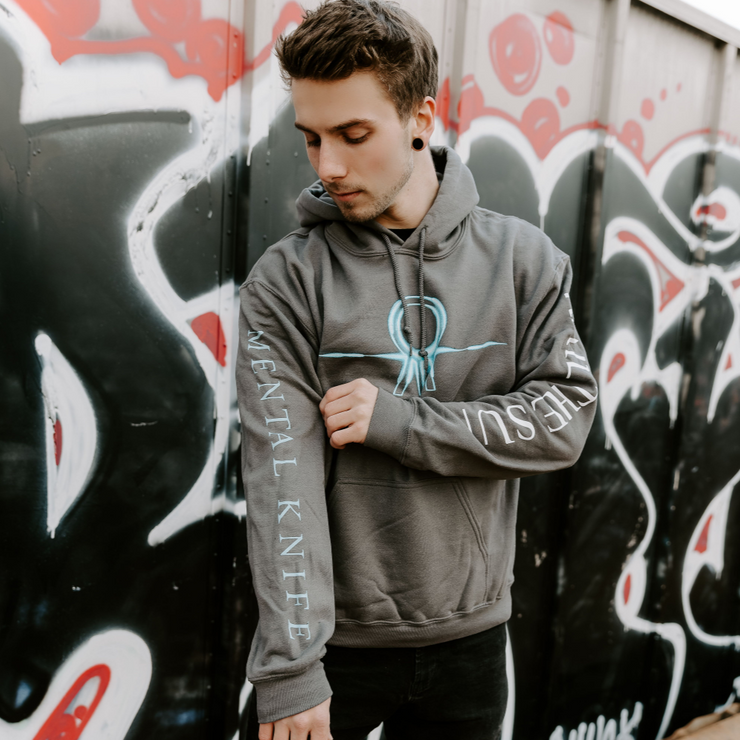 Someone wearing a Charcoal colored hoodie with the Hail The Sun logo across the chest in blue (circle with three lines coming out the bottom. On one sleeve, there is white text that says HAIL THE SUN, and on the other there is blue text that says MENTAL KNIFE. There is a graffitied storage container in the background.