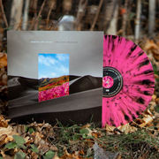 The background of this album is a gloomy black and white dune landscape. In the middle of the album cover is a rectangle with a clear blue sky with green hills and pink flowers. Above the rectangle reads ARMOR FOR SLEEP in white small letters and nect to it reads THE RAIN MUSEUM in black small letters. The LP is pink with black streak patterns.