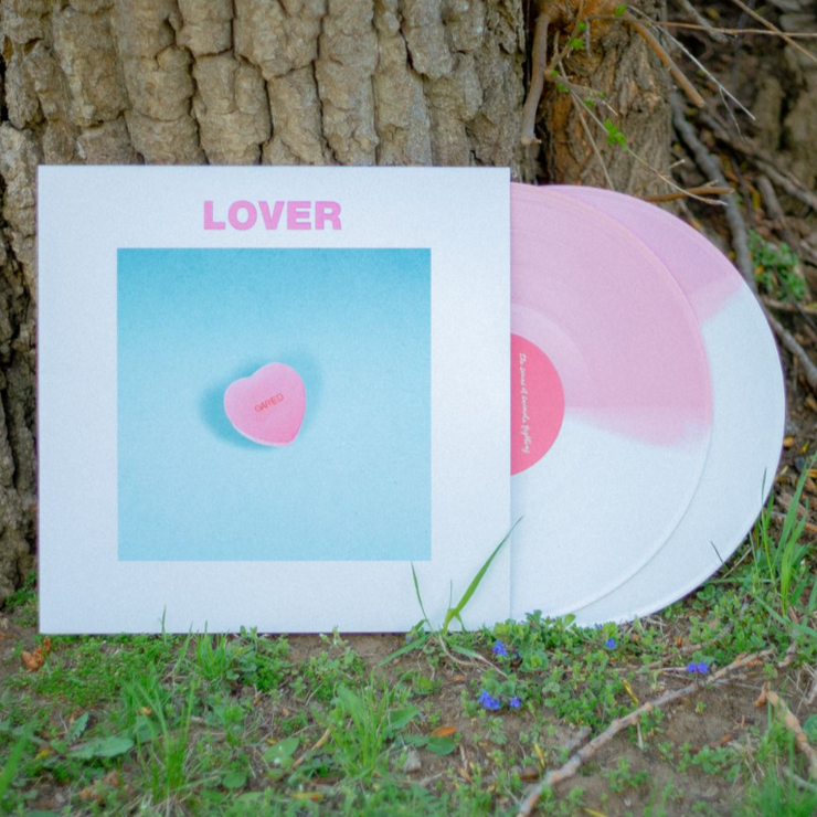 Vinyl cover with a picture of a heart candy and the text LOVER written above it. The two vinyl that are sticking out are white/Pink colored. It is all leaning against a tree on a spring day.