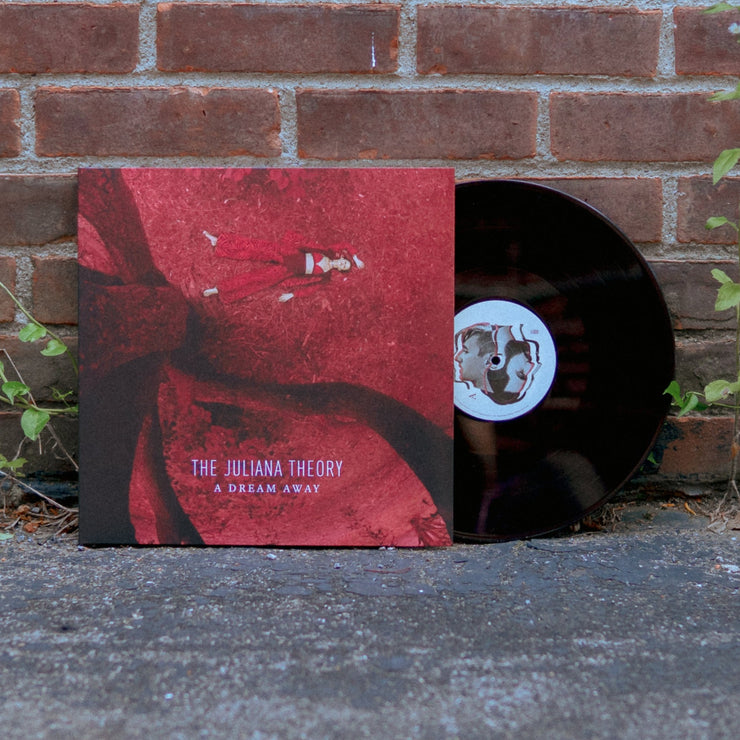 Black vinyl record coming out of vinyl jacket. Art on vinyl jacket is a girl laying on red ground next to the trunk of a tree. Photo is taken in an aerial above view. Text at the bottom of the vinyl jacket says THE JULIANA THEORY A DREAM AWAY. The vinyl is leaning on a brick wall. 