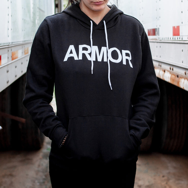 Black pullover hoodie with ARMOR written on the chest in large white font. On the bottom right sleeve is a small threaded figure. An individual is pictured wearing the hoodie between two large white trucks.