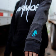 Black pullover hoodie with ARMOR written on the chest in large white font. On the bottom right sleeve is a small threaded figure. An individual is pictured wearing the hoodie between two large white trucks. Zoomed in picture of the threaded figure.