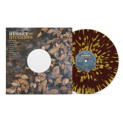 Vinyl jacket with an image of dead leaves on the ground and a hole in the center for the vinyl to show through. In the top left corner there is text that says HUSSEY HITCHENS, in white and orange coloring. Below that is a track list for the vinyl. To the right is the vinyl, which is burgundy with gold splatter.