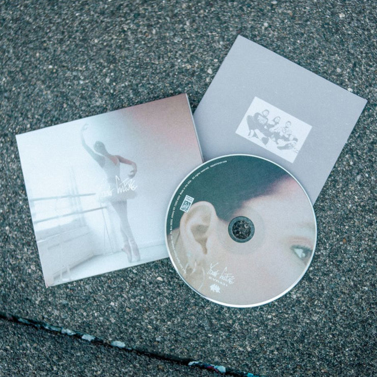 Square CD that says YOUNG CULTURE in the center in scratchy white font. The album art is an image of a ballerina dancing in a white room. There is a disc laying on top of the cover, which is an image of a close up of a woman's face. The bottom of the disc has text that says YOUNG CULTURE. Both are laying on top of asphalt.
