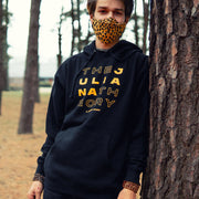 Black hoodie with "THE JULIANA THEORY" written in orange lettering in rows of four. The word 'Juliana' is a solid orange while "The" and "Theory" are black but outlined in orange. An individual is pictured wearing the hoodie, wearing a face mask and leaning up against a tree.
