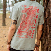 Back of sand colored short sleeve shirt with a large vertical rectangle filled with a red leaves design. To the right of the design is red text that says THE JULIANA THEORY. An individual is modeling the shirt, leaning up against a tree and there is a forest in the background.