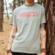 Sand colored short sleeve shirt with THE JULIANA THEORY written in red font across the chest. Below the text, there is a rectangle with a red design of leaves inside it. an individual is modeling the short, leaning up against a tree with a forest in the background.