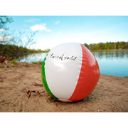 Red, blue, yellow, green, and white beach ball with "bars of gold" written in lowercase black cursive on the white side. Ball is on the sand on the bank of a body of water.