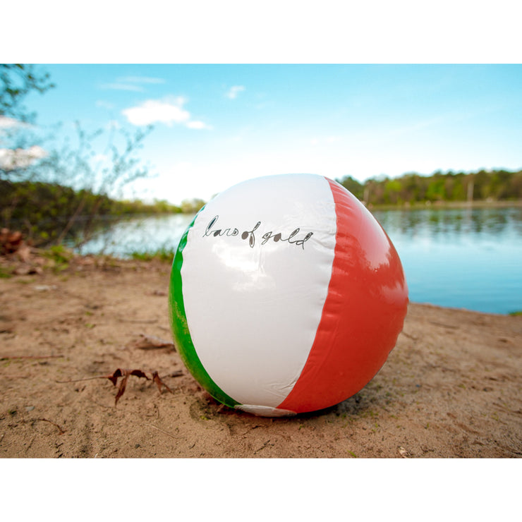 Red, blue, yellow, green, and white beach ball with "bars of gold" written in lowercase black cursive on the white side. Ball is on the sand on the bank of a body of water.