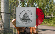 Vinyl jacket with a faceless person in a robe sitting with their legs crossed. Around them is a tree branch sprouting from one of their hands. Peeking out of the jacket is a red and black splattered vinyl. An individual is holding up both, standing next to a fence with trees and grass in the background.