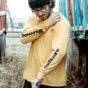 Mustard yellow long sleeve shirt with a small black globe in the top right. Both arms have vertical writing spelling "young culture" each word on opposite arms. An individual is wearing the long sleeve shirt, standing between two trucks in tall grass.