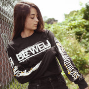 Black long sleeve shirt with BE WELL written in large white font across the chest. BE WELL is also written down one sleeve, and THE WEIGHT AND THE COST is written down the other. An individual is modeling the shirt and standing next to a fence with trees in the background.