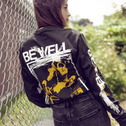 Back of black long sleeve shirt with BE WELL written in large font. Below that there is a drawing of someone playing guitar. Below the image is more white text that says THE WEIGHT AND THE COST. An individual is modeling the shirt and standing next to a fence with trees in the background.  