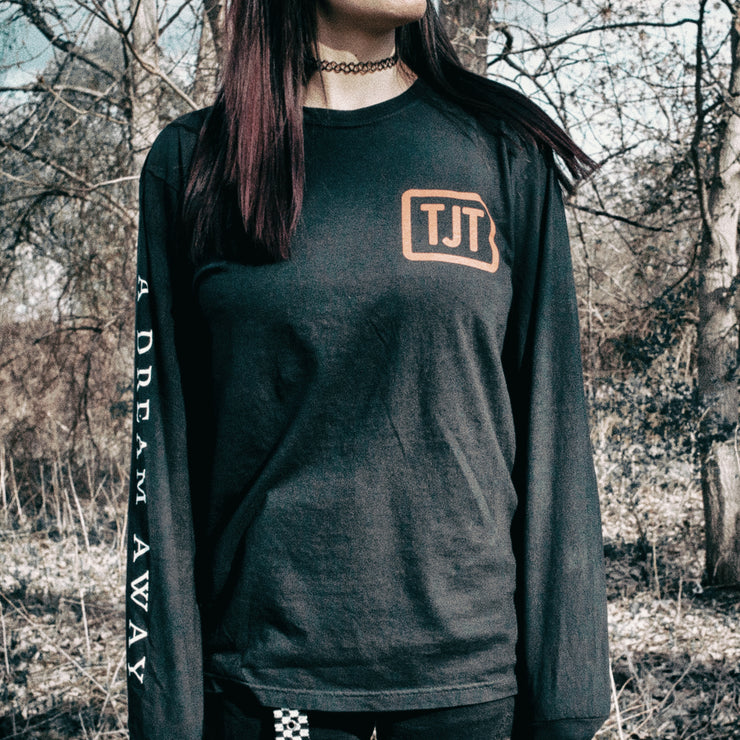 Black long sleeve shirt with a red box in the top corner with red text inside that says TJT. On one sleeve there is white text that says A DREAM AWAY. An individual is modeling the shirt and standing in the woods in front of dead trees.