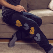 A person is sitting on a couch wearing black sweatpants with yellow and gray broken hearts running down the side of them. 