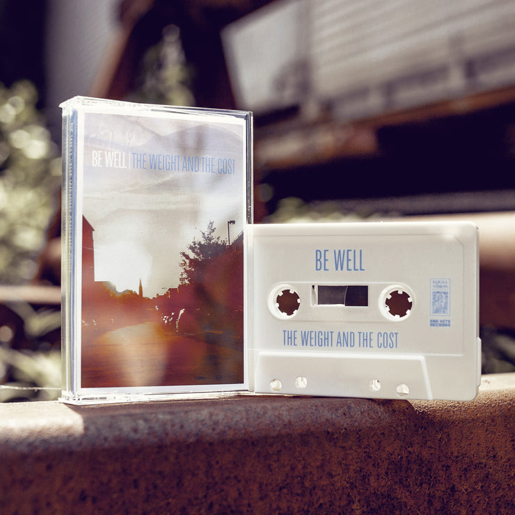 Cassette tape with white text that says BE WELL. Next to that there is blue text that says THE WEIGHT AND THE COST. The album artwork is an image taken down a street while the sun is setting. There are cars lining the street as well as trees and a street light. There is a white cassette tape next to it, with BE WELL and THE WEIGHT AND THE COST written in blue text. Both are standing up on a rusted surface.